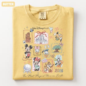 May include: Butter-colored t-shirt featuring a Walt Disney World graphic. The design includes illustrations of Mickey Mouse, Minnie Mouse, Donald Duck, and other characters, along with the text "The Most Magical Place on Earth" and the year 2026.