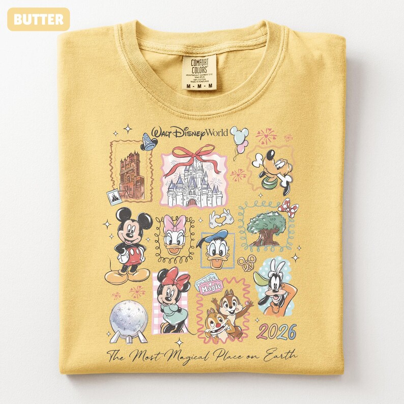 May include: Butter-colored t-shirt featuring a Walt Disney World graphic. The design includes illustrations of Mickey Mouse, Minnie Mouse, Donald Duck, and other characters, along with the text "The Most Magical Place on Earth" and the year 2026.