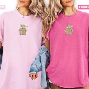 May include: Two short-sleeved t-shirts, one light pink and one bright pink, each featuring an embroidered image of a green character in a tan coat. The light pink shirt has the word "Blossom" above it. The bright pink shirt has the word "Crunchberry" above it.