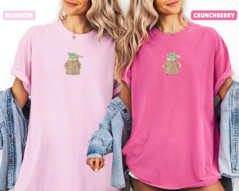 May include: Two short-sleeved t-shirts, one light pink and one bright pink, each featuring an embroidered image of a green character in a tan coat. The light pink shirt has the word "Blossom" above it. The bright pink shirt has the word "Crunchberry" above it.