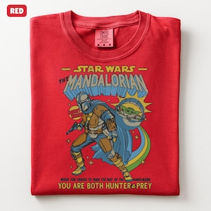 以下が含まれることがあります： スターウォーズのマンダロリアンのグラフィックが特徴の赤いTシャツ。デザインにはマンダロリアン、ベビーヨーダのようなキャラクター、そして「マンダロリアン」と「あなたはハンターと獲物の両方である」というテキストが含まれています。