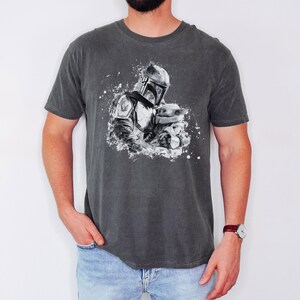 Op de afbeelding: Donkergrijs t-shirt met een zwart-witte afbeelding van een Mandalorian-personage dat een klein wezen vasthoudt. Het ontwerp heeft een spatteffect. Het shirt heeft korte mouwen en een ronde hals.