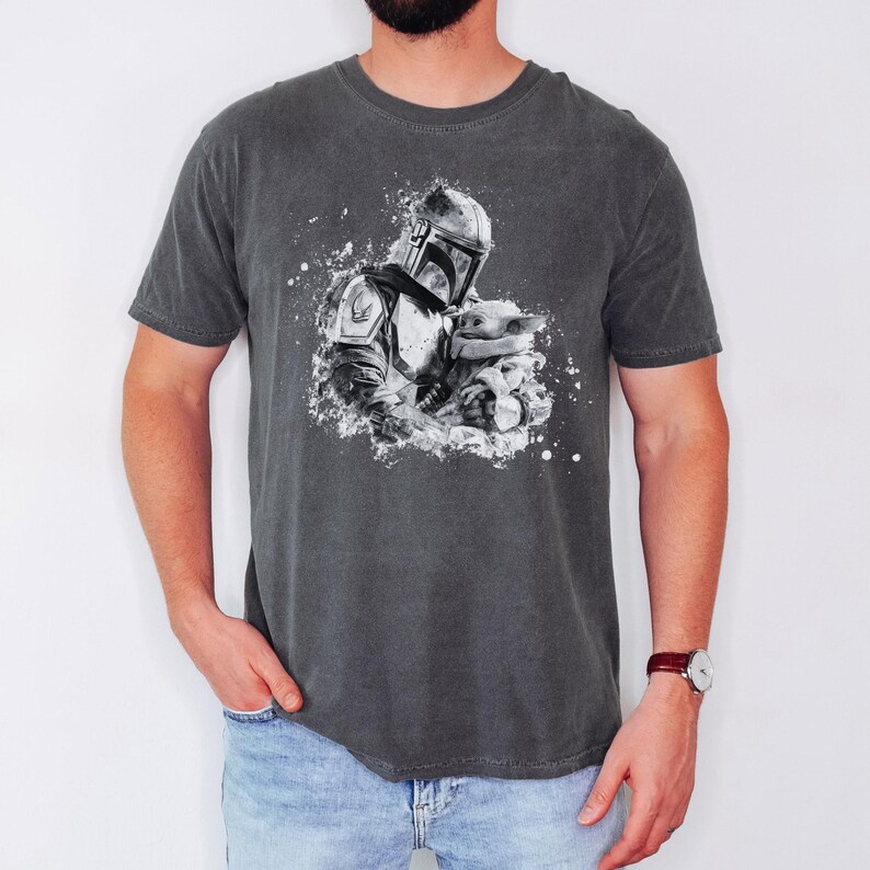 Op de afbeelding: Donkergrijs t-shirt met een zwart-witte afbeelding van een Mandalorian-personage dat een klein wezen vasthoudt. Het ontwerp heeft een spatteffect. Het shirt heeft korte mouwen en een ronde hals.