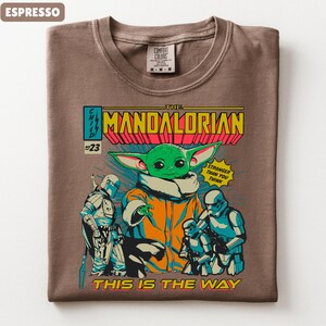 May include: Espresso-colored t-shirt featuring a comic book-style graphic of The Mandalorian. The design includes the character Grogu, Stormtroopers, and the text "THIS IS THE WAY". The shirt has a crew neck and a Comfort Colors tag.