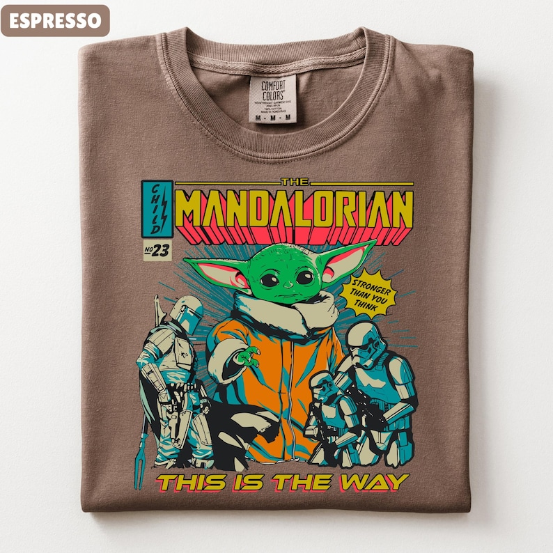 May include: Espresso-colored t-shirt featuring a comic book-style graphic of The Mandalorian. The design includes the character Grogu, Stormtroopers, and the text "THIS IS THE WAY". The shirt has a crew neck and a Comfort Colors tag.