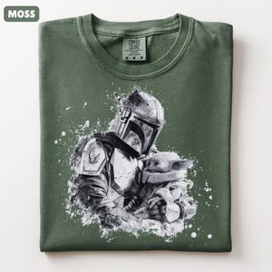 Op de afbeelding: Mosgroen t-shirt met een zwart-witte afbeelding van een Mandalorian-personage dat een klein wezen vasthoudt. De afbeelding heeft een distressed, splatter-paint effect. Het shirt heeft een ronde hals en een label "Comfort Colors".