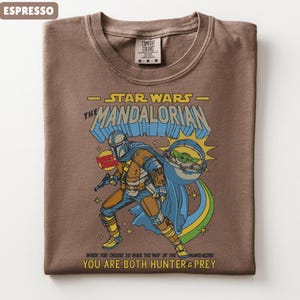 以下が含まれることがあります： スターウォーズのマンダロリアンが描かれたエスプレッソカラーのTシャツ。デザインにはマンダロリアン、グローグー、そして「YOU ARE BOTH HUNTER & PREY」というテキストが含まれています。このシャツはクルーネックで、Comfort Colorsのタグが付いています。
