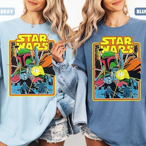 May include: Light blue and blue jean Star Wars graphic t-shirts. Each shirt features a colorful graphic of Boba Fett and other characters, with the Star Wars logo in yellow and orange. The shirts have short sleeves and a relaxed fit.