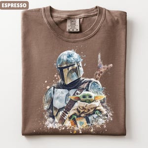 Puede incluir: Camiseta color espresso con una ilustraci&oacute;n estilo acuarela de The Mandalorian y Grogu. El dise&ntilde;o incluye el casco y la armadura del Mandaloriano, y Grogu envuelto en una manta. La camiseta tiene cuello redondo y una etiqueta Comfort Colors.