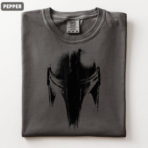 May include: A folded pepper-coloured t-shirt with a black Mandalorian helmet graphic. The design is prominently displayed. The tag reads "Comfort Colors" with size details. A relaxed-fit tee, ideal for fans of the series.