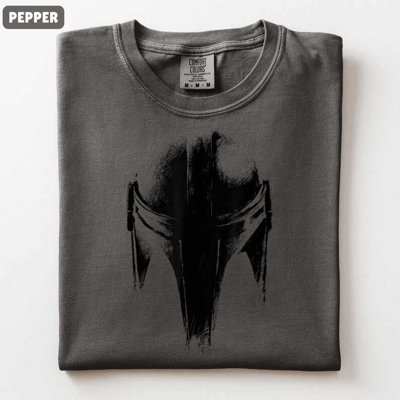 May include: A folded pepper-coloured t-shirt with a black Mandalorian helmet graphic. The design is prominently displayed. The tag reads "Comfort Colors" with size details. A relaxed-fit tee, ideal for fans of the series.