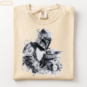Op de afbeelding: Ivoor kleurig T-shirt met een zwart-wit afbeelding van een Mandalorian karakter met Grogu. Het shirt heeft een ronde hals en een label met "Comfort Colors". De afbeelding heeft een aquareleffect.