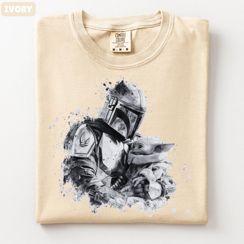 Op de afbeelding: Ivoor kleurig T-shirt met een zwart-wit afbeelding van een Mandalorian karakter met Grogu. Het shirt heeft een ronde hals en een label met "Comfort Colors". De afbeelding heeft een aquareleffect.