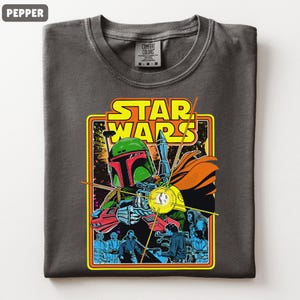 May include: Pepper-colored t-shirt featuring a Star Wars graphic. The graphic includes the text "STAR WARS" in yellow and orange, and an illustration of Boba Fett with a blaster. The shirt is a Comfort Colors brand.