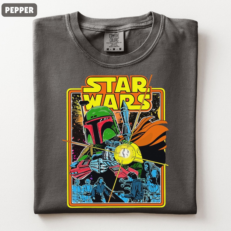May include: Pepper-colored t-shirt featuring a Star Wars graphic. The graphic includes the text "STAR WARS" in yellow and orange, and an illustration of Boba Fett with a blaster. The shirt is a Comfort Colors brand.