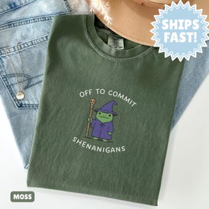 Puede incluir: Camiseta verde musgo con el texto "OFF TO COMMIT SHENANIGANS" y una rana de dibujos animados con sombrero y túnica de mago. La rana sostiene un bastón. La camiseta está doblada sobre unos vaqueros azules.