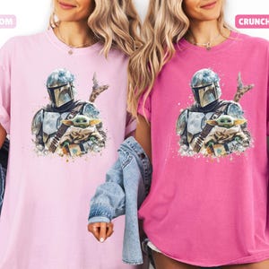 Puede incluir: Dos camisetas, una rosa claro y otra magenta, cada una con una ilustraci&oacute;n en acuarela de un personaje mandaloriano y Grogu. La camiseta rosa claro tiene la palabra "BLOSSOM" en un cuadro rosa. La camiseta magenta tiene la palabra "CRUNCHBERRY" en un cuadro rosa.