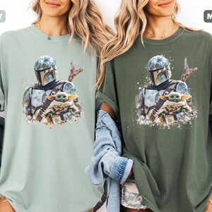 Puede incluir: Dos camisetas, una en color verde claro y otra en verde oscuro, cada una con una ilustraci&oacute;n al estilo acuarela. La ilustraci&oacute;n representa a un personaje con casco sosteniendo una peque&ntilde;a criatura verde. Las camisetas son informales y tienen las mangas dobladas.