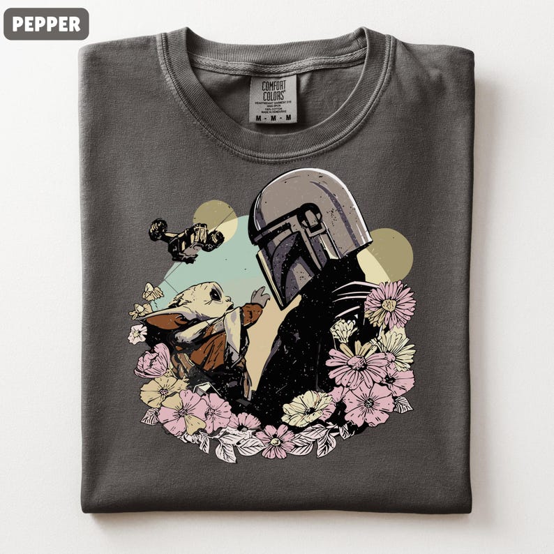 May include: Pepper-colored t-shirt featuring a graphic of a Mandalorian helmet and Grogu surrounded by flowers. A ship flies in the background. The shirt is a Comfort Colors brand.