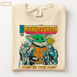 May include: Ivory-colored t-shirt featuring a comic book-style graphic of The Mandalorian. The design includes the Child (Grogu), Stormtroopers, and the text "The Mandalorian" and "This is the Way."