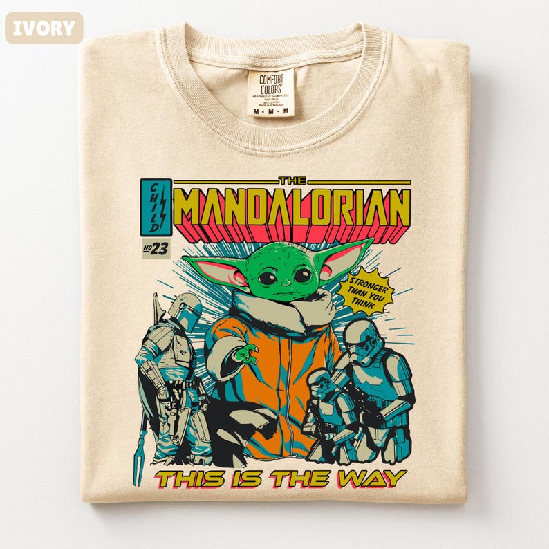 May include: Ivory-colored t-shirt featuring a comic book-style graphic of The Mandalorian. The design includes the Child (Grogu), Stormtroopers, and the text "The Mandalorian" and "This is the Way."