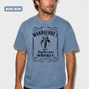 Puede incluir: Camiseta azul vaquera con el texto "MANDALORE'S Mythosaur WHISKEY" en negro. La camiseta presenta un gr&aacute;fico de una calavera y el texto "70cl" y "40%" en la parte inferior. La camiseta es de manga corta.