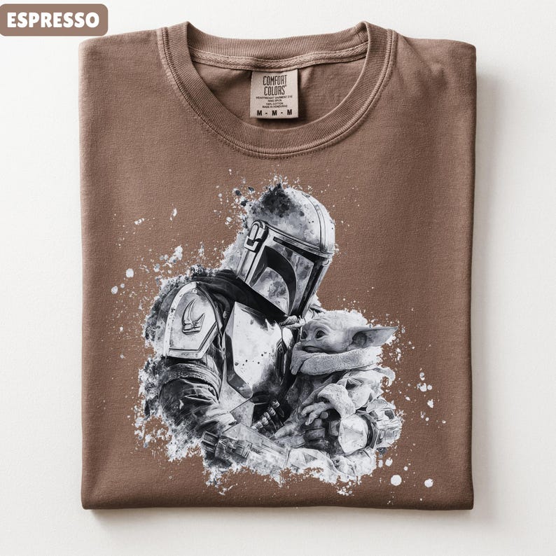 Op de afbeelding: Espresso-kleurig T-shirt met een zwart-witte afbeelding van een Mandalorian-personage dat een klein wezen vasthoudt. Het shirt heeft een ronde hals en een Comfort Colors-label.