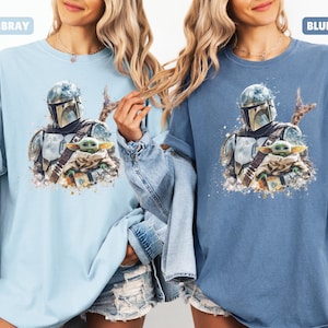 Puede incluir: Dos camisetas, una azul claro y otra azul, cada una con una ilustraci&oacute;n al estilo acuarela de The Mandalorian y Grogu. La camiseta azul claro est&aacute; etiquetada como "CHAMBRAY" y la azul como "BLUE JEAN".