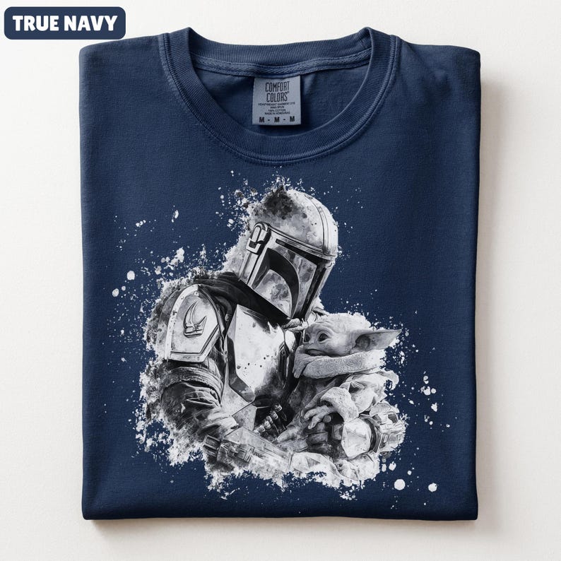 Op de afbeelding: Marineblauw T-shirt met een zwart-witte afbeelding van een Mandalorian-personage dat een klein wezen vasthoudt. Het ontwerp heeft een spatteffect. Het shirt heeft een ronde hals en een label "Comfort Colors".