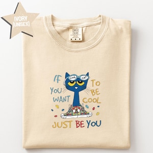 May include: An ivory t-shirt featuring a graphic of a blue cat wearing sunglasses and colorful patterned sneakers. The text reads: If you want to be cool just be you. The cat is surrounded by small puzzle piece icons in various colors. The shirt has a crew neck.