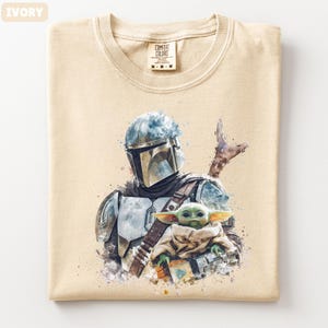 Puede incluir: Camiseta de color marfil con una ilustraci&oacute;n en acuarela de un personaje mandaloriano y Grogu. El dise&ntilde;o incluye un casco, una armadura y una peque&ntilde;a criatura verde. La camiseta tiene cuello redondo y una etiqueta que dice "Comfort Colors".