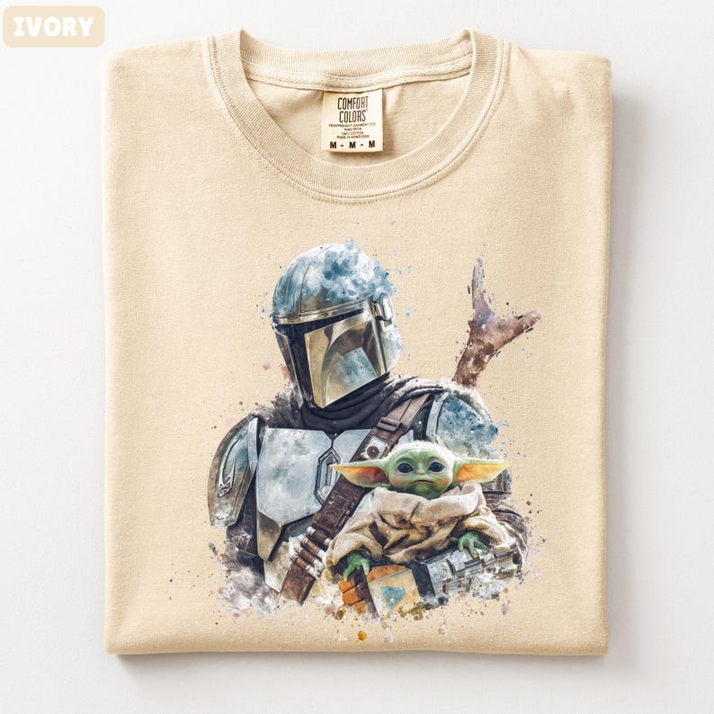 Puede incluir: Camiseta de color marfil con una ilustraci&oacute;n en acuarela de un personaje mandaloriano y Grogu. El dise&ntilde;o incluye un casco, una armadura y una peque&ntilde;a criatura verde. La camiseta tiene cuello redondo y una etiqueta que dice "Comfort Colors".