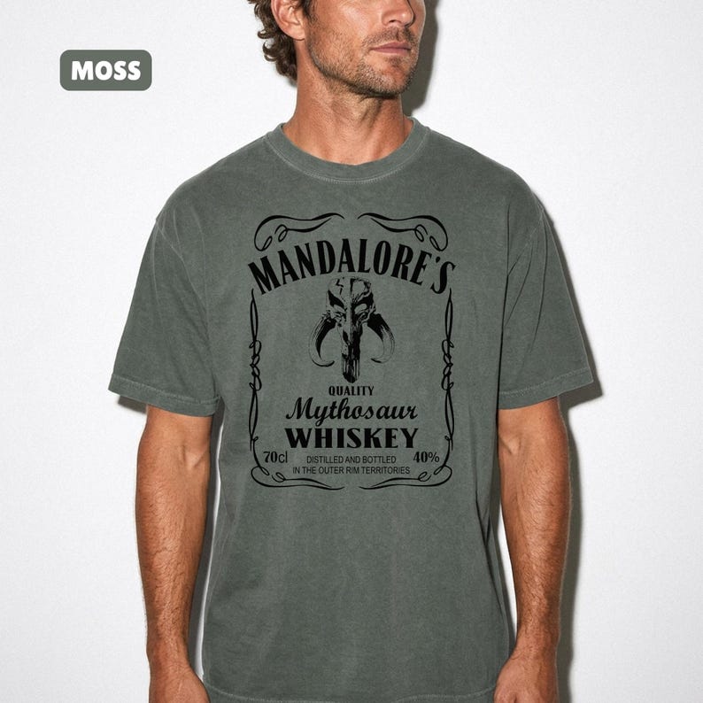 Puede incluir: Camiseta verde musgo con un gr&aacute;fico negro que se asemeja a una etiqueta de whisky. La etiqueta dice "MANDALORE'S" encima de una calavera de Mythosaur, con "Mythosaur WHISKEY" debajo. La camiseta tambi&eacute;n indica 70cl y 40% de alcohol.