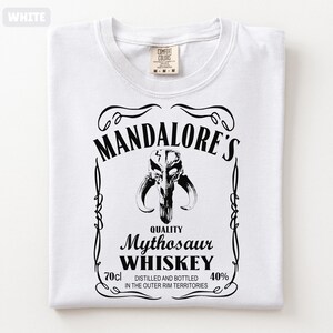 Puede incluir: Camiseta blanca con un dise&ntilde;o gr&aacute;fico negro. El dise&ntilde;o se asemeja a una etiqueta de whisky y presenta el texto "MANDALORE'S Mythosaur WHISKEY" junto con una imagen de calavera. La camiseta tambi&eacute;n incluye el texto "70cl" y "40%."