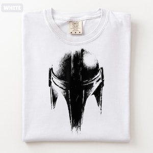 May include: White t-shirt with a black illustration of a Mandalorian helmet. The graphic is a detailed, artistic rendering. The shirt is folded and has a Comfort Colors label.
