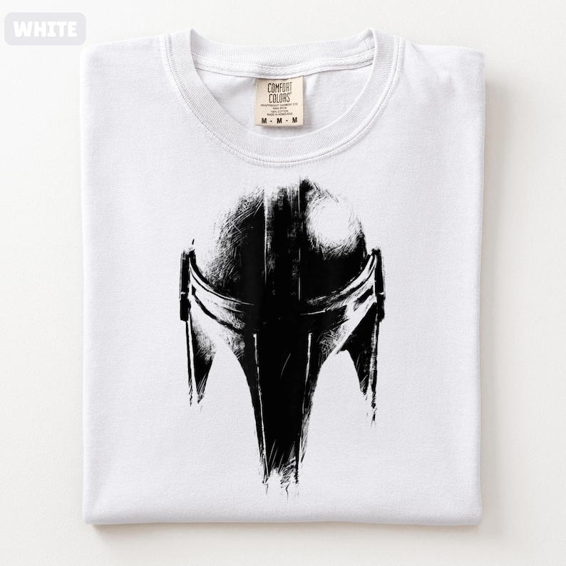 May include: White t-shirt with a black illustration of a Mandalorian helmet. The graphic is a detailed, artistic rendering. The shirt is folded and has a Comfort Colors label.