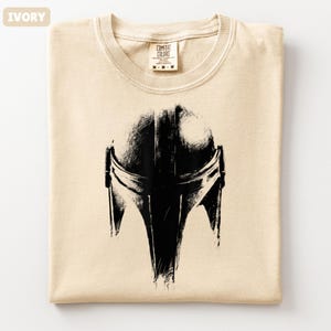 May include: Ivory t-shirt featuring a black Mandalorian helmet graphic. The shirt is folded, with the Comfort Colors label at the neck. The design is a detailed, stylized illustration of the helmet.