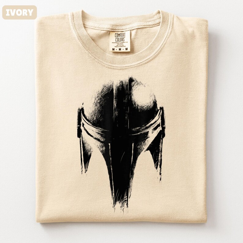 May include: Ivory t-shirt featuring a black Mandalorian helmet graphic. The shirt is folded, with the Comfort Colors label at the neck. The design is a detailed, stylized illustration of the helmet.