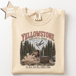May include: An ivory t-shirt featuring a graphic of a person being tossed into the air by a bison in a forest setting. The text reads Yellowstone National Park and Do Not Pet The Fluffy Cows. The shirt has a crew neck and a Comfort Colors brand label.