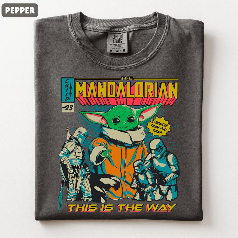 May include: Pepper-colored t-shirt featuring a comic book-style graphic of The Mandalorian. The design includes the Child (Grogu), Stormtroopers, and the text "THIS IS THE WAY". The shirt has a crew neck and a Comfort Colors tag.