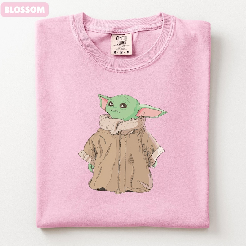 May include: A light pink t-shirt featuring a graphic of a green alien-like character wearing a brown coat. The shirt has a crew neck and a tag that reads "Comfort Colors". The word "Blossom" is in the top left corner.