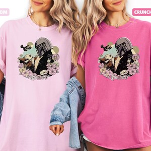 May include: Two t-shirts, one light pink and one hot pink, featuring a graphic of a Mandalorian character and a small creature surrounded by flowers. The light pink shirt has the word "BLOSSOM" and the hot pink shirt has the word "CRUNCHBERRY".