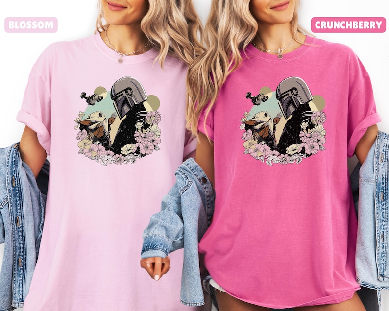 May include: Two t-shirts, one light pink and one hot pink, featuring a graphic of a Mandalorian character and a small creature surrounded by flowers. The light pink shirt has the word "BLOSSOM" and the hot pink shirt has the word "CRUNCHBERRY".