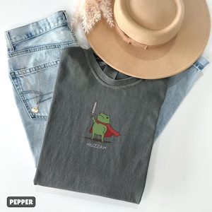 May include: A gray t-shirt with a cartoon frog wearing a red cape and holding a sword, with the word "Huzzah" below. The shirt is paired with light blue jeans and a tan hat.