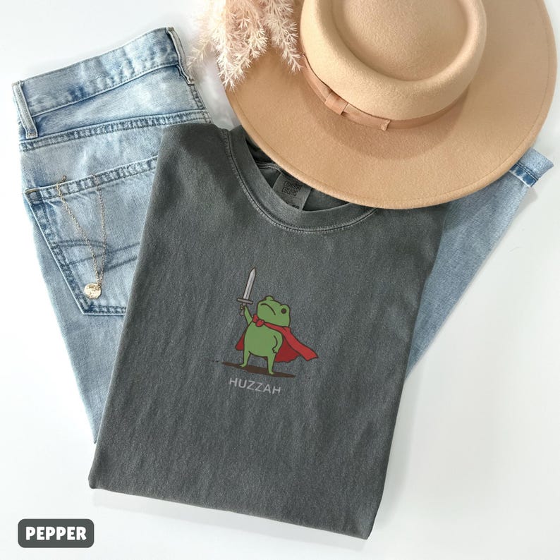 May include: A gray t-shirt with a cartoon frog wearing a red cape and holding a sword, with the word "Huzzah" below. The shirt is paired with light blue jeans and a tan hat.