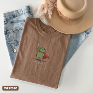 May include: Espresso-colored t-shirt featuring a cartoon frog with a sword and cape, with the word "Huzzah" below. The shirt is paired with light blue jeans and a tan hat.