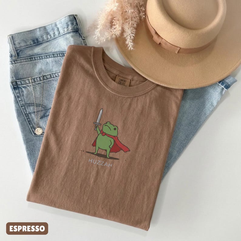 May include: Espresso-colored t-shirt featuring a cartoon frog with a sword and cape, with the word "Huzzah" below. The shirt is paired with light blue jeans and a tan hat.