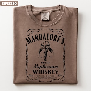 Puede incluir: Camiseta color espresso con un dise&ntilde;o gr&aacute;fico negro que se asemeja a una etiqueta de whisky. El dise&ntilde;o incluye el texto "MANDALORE'S" encima de una calavera de Mythosaur, con "Mythosaur WHISKEY" debajo. La camiseta tambi&eacute;n tiene una etiqueta "COMFORT COLORS".