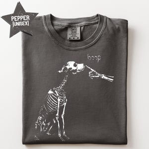 以下が含まれることがあります： チャコールグレーのTシャツで、骸骨の指が犬の骸骨の鼻先を触っている白いグラフィックがプリントされています。犬の頭の上には「boop」という文字があります。クルーネックで、Comfort Colorsのブランドラベルが付いています。