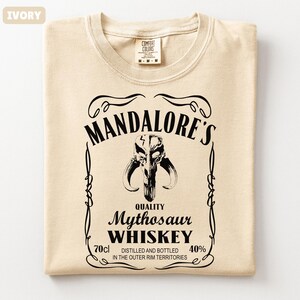Puede incluir: Camiseta de color marfil con un gr&aacute;fico negro que se asemeja a una etiqueta de whisky. La etiqueta dice "MANDALORE'S Mythosaur WHISKEY" con un emblema de calavera. La camiseta tambi&eacute;n tiene el texto "70cl" y "40%" en la etiqueta.